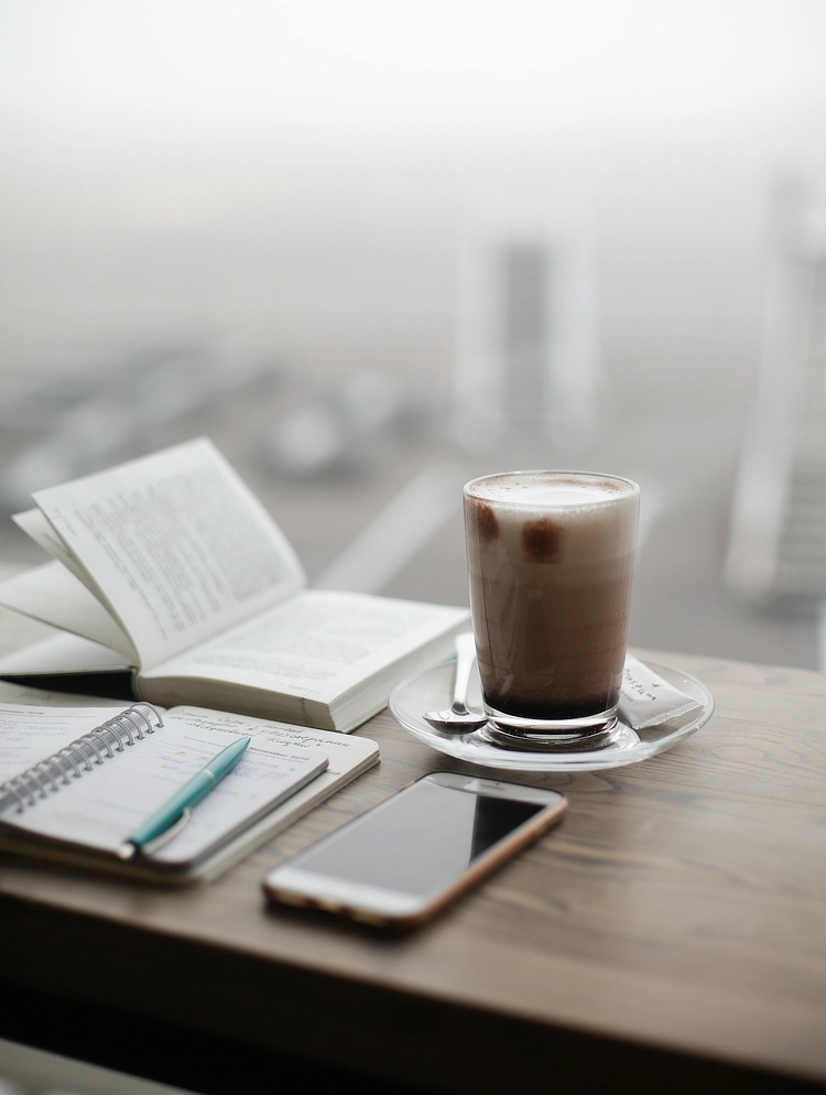 A wooden table top with a phone, coffee, and notebooks